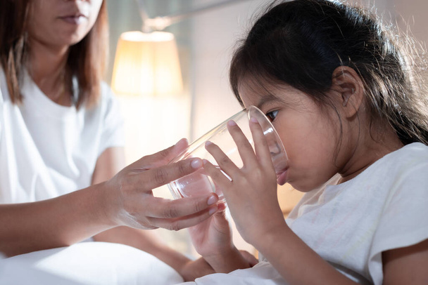 Madre asiática dando vaso de agua a niña enferma después de comer medicina en el dormitorio. Concepto de niño enfermo y cuidado de salud. - Foto, imagen