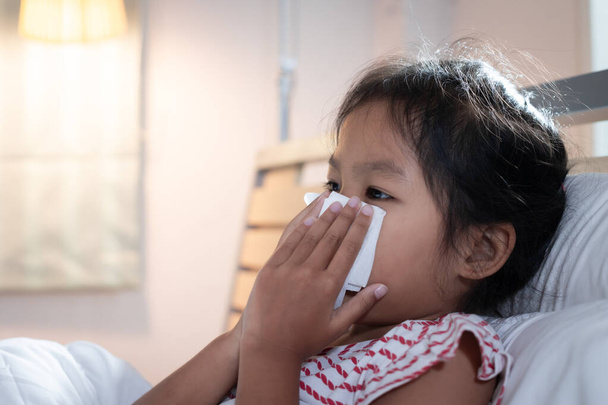 Азиатская девочка заболевает, кашляет и чихает. Она вытирает нос бумажными салфетками. Больной ребенок и концепция здравоохранения. - Фото, изображение