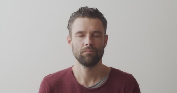 Közelről a férfi arc elől jóga gyakorlatokat végez beltérben, fehér háttérrel. Szakállas férfi csukott szemmel pránajámát gyakorol természetes fény lassított felvételen. Egészséges test egészségügyi koncepció - Felvétel, videó