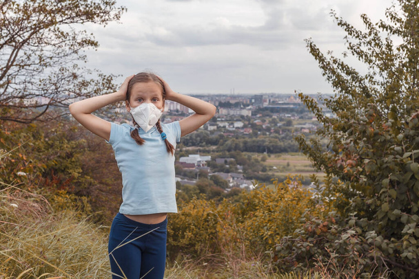 Little girl in a T-shirt in a medical mask on his face. The child grabbed his head with his hands. Girl on a background of the city - Photo, Image