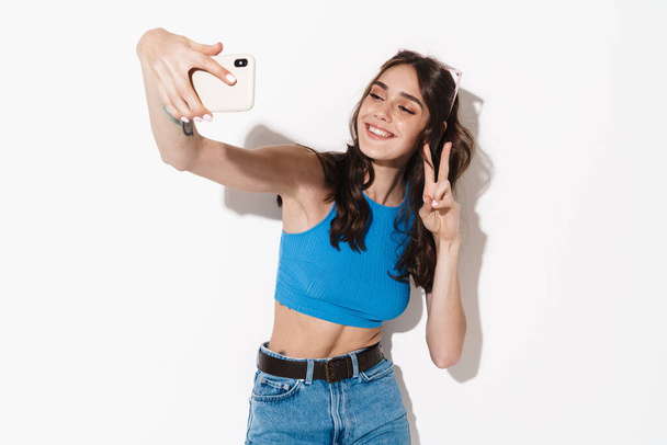 Image of happy woman making peace gesture while taking selfie on cellphone isolated over white background - Zdjęcie, obraz