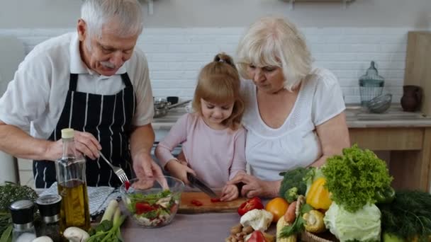 Senior couple in kitchen teaching granddaughter child how to cook, chopping pepper with knife - Footage, Video