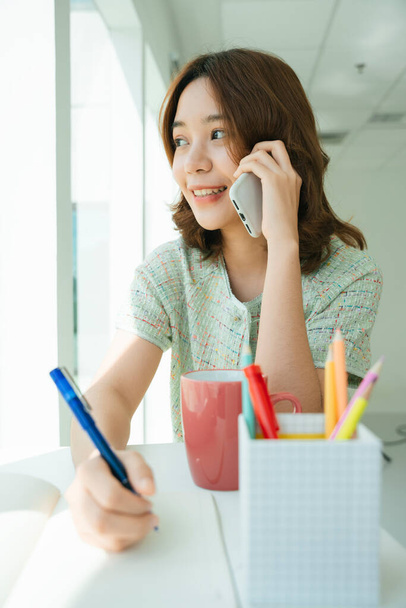 Portrét mladé asijské ženy napsat knihu a mluvit po telefonu. - Fotografie, Obrázek