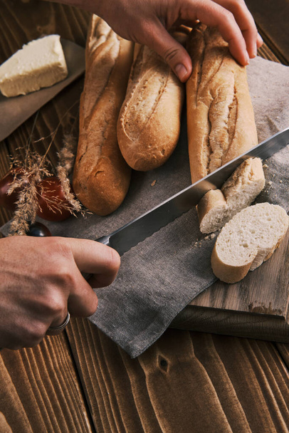 Cerrar las manos rebanando baguette francesa fresca en un paño de lino sobre una mesa de madera - Foto, Imagen