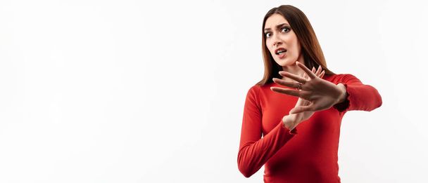 Image of dissatisfied girl with long chestnut hair in casual red sweater doing disgust face because aversion reaction with hands raised. Studio shot, white background. Facial expression concept - Photo, Image