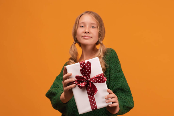 Kép a bájos szőke lány 12-14 éves meleg zöld pulóver mosolygó és gazdaság jelen doboz piros íj. Stúdiófelvétel, sárga háttér. Szilveszter Női Nap születésnap ünnep koncepció - Fotó, kép