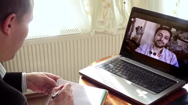 Homme adulte âgé travaillant à la maison prenant des notes dans un appel vidéo qu'il a l'aide de son ordinateur portable avec un collègue plus jeune - Séquence, vidéo