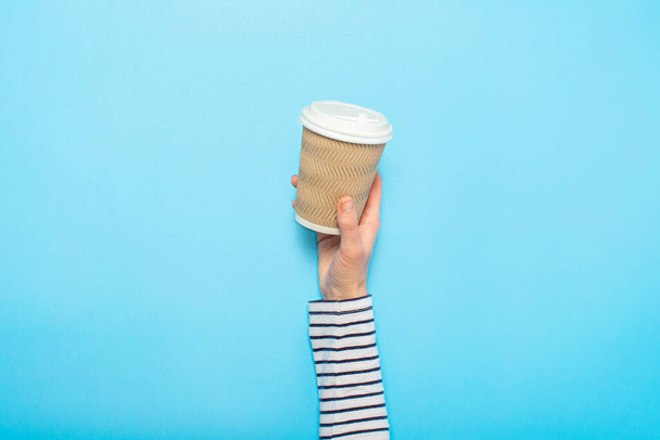 Female hand holds a paper cup with coffee on a blue background. Concept of coffee, a warming drink, breakfast. Banner Flat lay, top view. - Фото, зображення