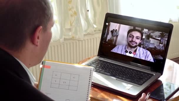 Middle aged man working from home taking notes in a video call he's having using his laptop with a younger colleague - Footage, Video
