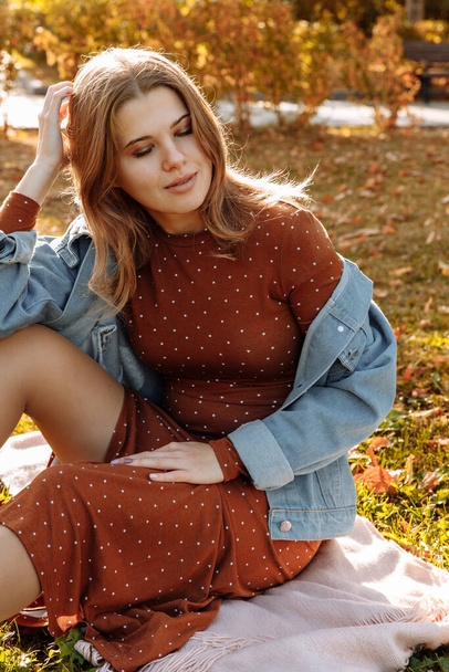 A young woman is sitting on the autumn ground in a polka-dot dress and jeans.  - Foto, Imagem