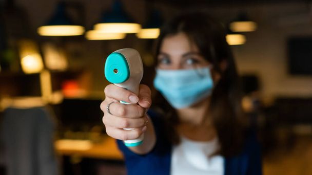 A female office worker in a mask holds a contactless electronic thermometer - Φωτογραφία, εικόνα