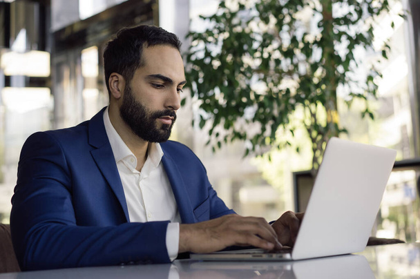 Young handsome arabic businessman planning project, typing on keyboard in modern office. Portrait of pensive middle eastern man wearing formal clothes, working online, sitting at workplace - 写真・画像