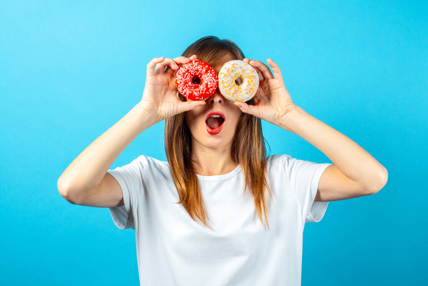 happy young girl in a white blank t-shirt, holds donuts in front of the eyes, on a blue background. Banner. Good mood, diet concept, bright colors. - Photo, Image