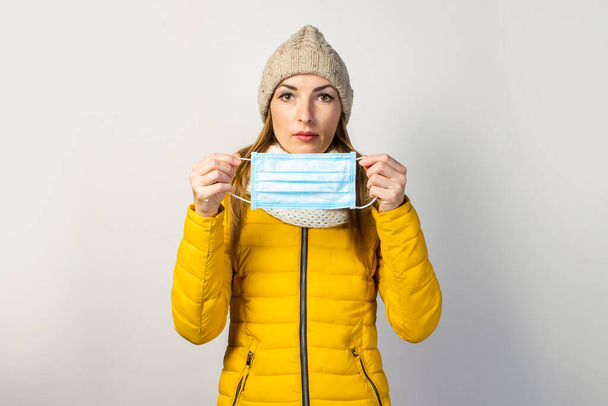 niña con una chaqueta y un sombrero amarillos, sostiene una máscara médica sobre un fondo claro. Concepto de resfriado común, virus, enfermedades infecciosas. - Foto, Imagen