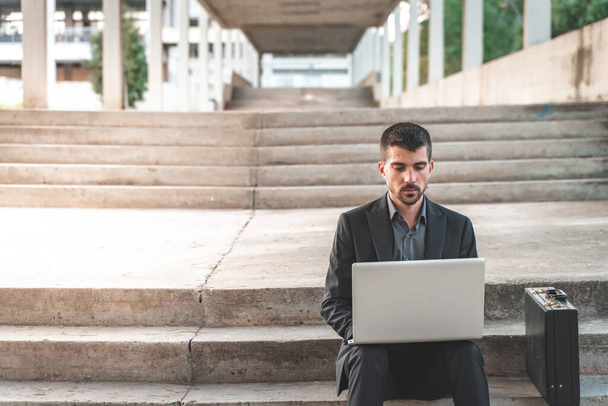 Handsome young businessman sitting on the stairs outdoors working with his laptop. Distance working. Isolated man on a suit. Copy space for text. - Valokuva, kuva