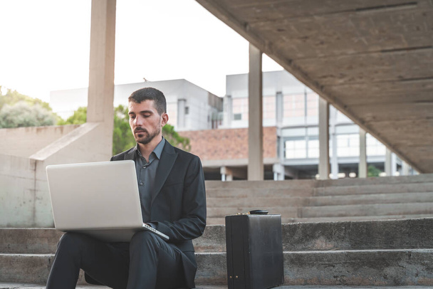 Handsome young businessman sitting on the stairs outdoors working with his laptop. Distance working. Isolated man on a suit. Copy space for text. - Foto, Bild
