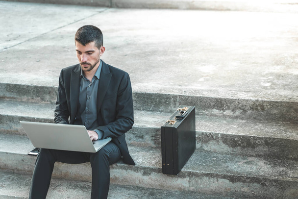 Handsome young businessman sitting on the stairs outdoors working with his laptop. Distance working. Isolated man on a suit. Copy space for text. - Foto, immagini