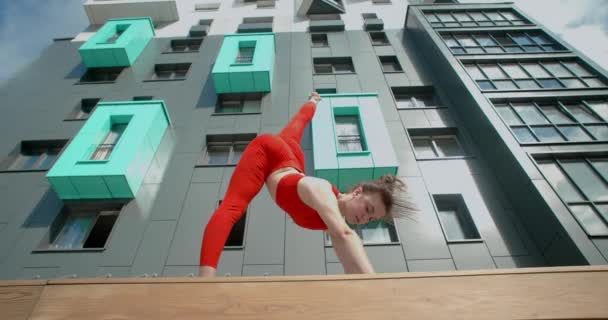 A vörös ruhás fiatal nő szalagos kallisztenikát végez a nappaliban, a tornász akrobatikus gyakorlatokat végez egy padon, városi környezetben fitnesz, 4k 120p Prores HQ - Felvétel, videó