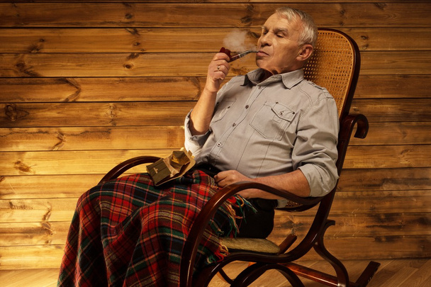 Homem sênior com cachimbo de fumar sentado na cadeira de balanço no interior de madeira caseira
 - Foto, Imagem