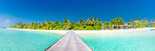 Удивительная панорама пляжа в Мальдиве. Элитные курортные виллы, мыс с причалом на острове Парадизе, пальмы над белым песком и голубым небом. Перфектный летний отдых, путешествия в отпуск - Фото, изображение