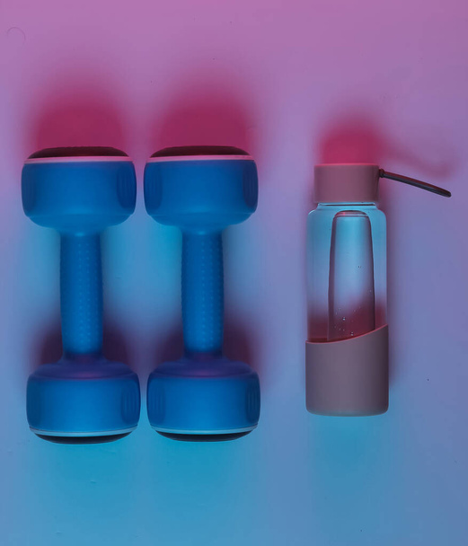 Fitness, sport concept. Dumbbells and a bottle of water. Neon pink blue night light.  - Фото, изображение