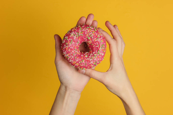 Female hands holding a glazed donut on yellow background. Sweet dessert. Top view. - 写真・画像
