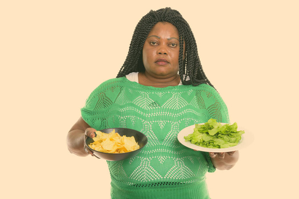Studio shot of fat black African woman holding bowl of potato chips and lettuce served on white plate - Valokuva, kuva