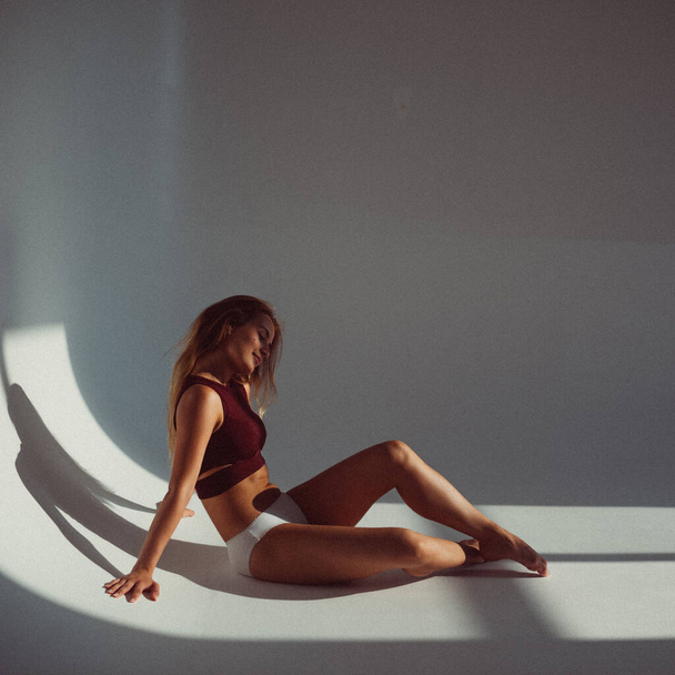 A young girl, with a plastic figure, licks on a white background in the studio, the sunlight from the window shines on her and creates a picture on the floor. she's wearing a sports top and white swimming trunks - Fotó, kép