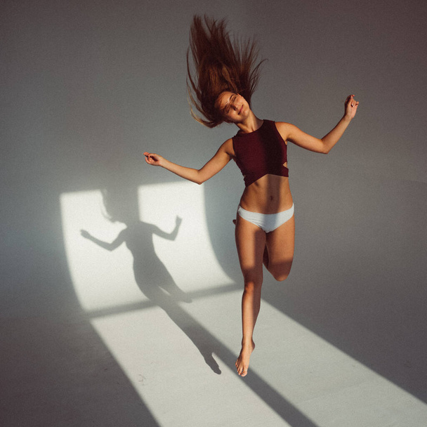 A young girl, with a plastic figure, jumps on a white background in the studio, the sunlight from the window shines on her and creates a picture on the floor. she's wearing a sports top and shorts - Zdjęcie, obraz