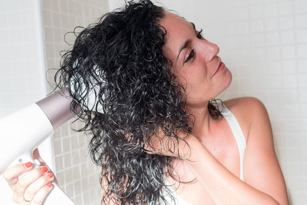 Eine junge Frau im Profil lächelt und kräuselt ihre Haare mit einem Haartrockner mit einer speziellen Düse zum Lockenwickeln. Trockner mit Diffusor. Pflege- und Schönheitskonzept. - Foto, Bild