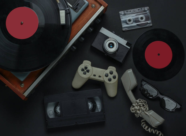 Flat lay retro media and entertainment. Vinyl record player with vinyl record, film camera, video cassette, audio cassette, gamepad, handset on a black background. 80s. Top view - Photo, Image