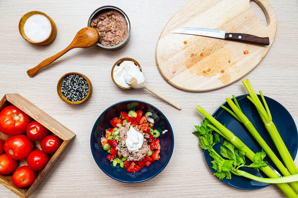 Ensalada con apio, tomate y atún. Ingredientes de ensalada en el proceso de cocción en una mesa de madera ligera vista superior - Foto, imagen