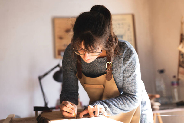 Молодая плотница рисует на куске древесины во время работы с деревом в столярной мастерской. Молодая работница деревообработки разрабатывает новую мебель. Концепция интерьера - Фото, изображение