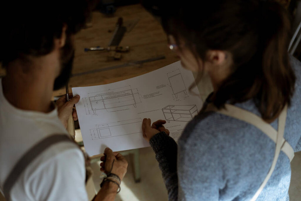 木の労働者の若いカップルは青写真シートの新しいプロダクト設計プロジェクトを勉強する。若い大工は一緒にインテリアデザインプロジェクトのための新しい家の家具を手作り. - 写真・画像
