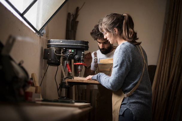 若い大工のカップルは、木材のためのドリルマシンを使用して小さな木の研究室で一緒に働いています。大工のワークショップで新しい家具を作るカップル。若い起業家は - 写真・画像