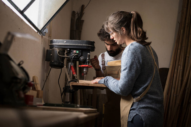A young couple of carpenter working together in a small wood laboratory using a drill machine for timber. Couple crafting new home furniture in a carpentry workshop. young entrepreneurs concept - Fotografie, Obrázek