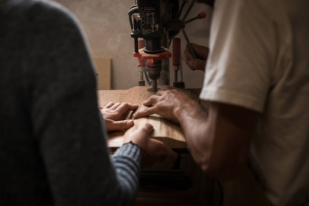 Ένα νεαρό ζευγάρι ξυλουργών που εργάζονται μαζί σε ένα μικρό εργαστήριο ξύλου χρησιμοποιώντας μια μηχανή γεώτρησης για ξυλεία. Ζευγάρι που κατασκευάζει νέα έπιπλα σπιτιού σε εργαστήριο ξυλουργικής. έννοια των νέων επιχειρηματιών - Φωτογραφία, εικόνα