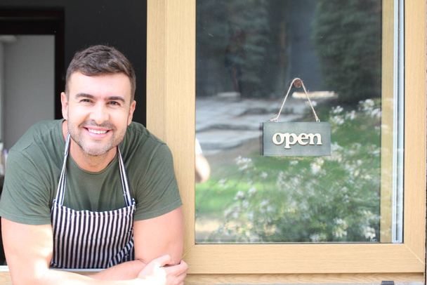 közeli kép egy jóképű fiatalemberről csíkos kötényben az üvegajtó előtt, nyitott táblával - Fotó, kép