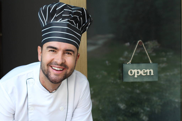 lähikuva muotokuva komea nuori kokki univormu ja hattu edessä ravintolan oven auki välilehti - Valokuva, kuva