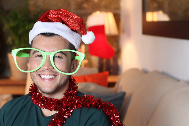 Porträt eines gutaussehenden jungen Mannes mit Weihnachtsmütze, der zu Hause während der Pandemie Weihnachten mit einer riesigen Brille feiert - Foto, Bild