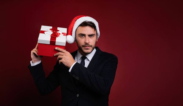 Young handsome caucasian guy in business suit and Santa hats stands on white background in studio and smilie Holds near the ear red gift box Portrait business person with Christmas mood Holiday banner - Zdjęcie, obraz