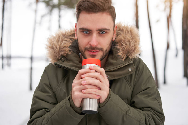 One Attractive Caucasian man stands winter forest  drink hot beverage thermos. Winter holiday leisure. Cold season. Portrait young handsome unshaved Modern hipster guy traveling walking in winter park - Photo, Image