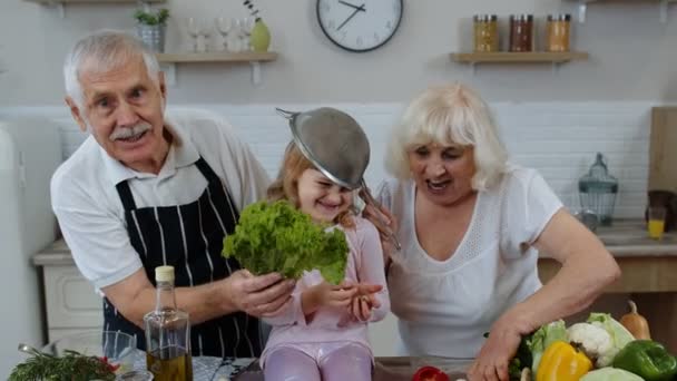 Мужчина и женщина старшего возраста с внучкой делают смешной танец со стрейнером и овощами дома - Кадры, видео