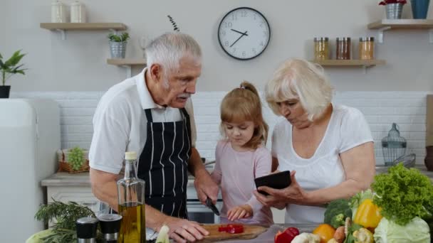 Senior grandparents couple with digital tablet and granddaughter cutting vegetables in kitchen - Footage, Video