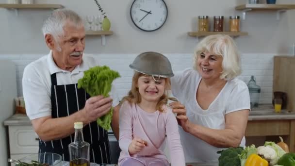 Femme âgée et homme avec petit-enfant fille qui batifolent avec passoire et légumes à la maison - Séquence, vidéo