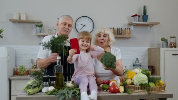 Blogger girl tomando fotos en el teléfono móvil con abuelos mayores en la cocina con verduras - Imágenes, Vídeo