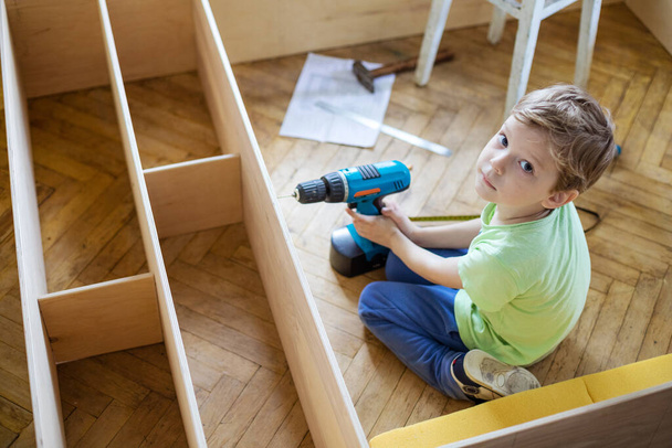 Niño con destornillador mirando hacia arriba mientras está sentado en el suelo en la estantería o estantería sin terminar - Foto, imagen