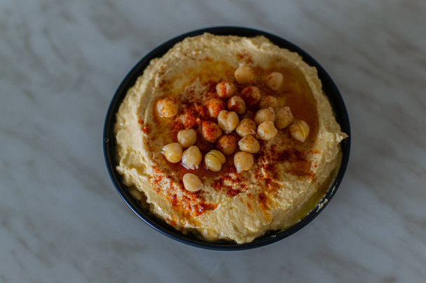 Creamy hummus with chickpeas, olive oil and red spices in dark bowl in vegetarian kitchen. Top view - Foto, Bild