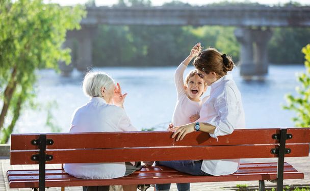 Vanhempi nainen, hänen aikuinen lapsenlapsensa ja lapsenlapsensa istuvat penkillä kesäpuistossa - Valokuva, kuva