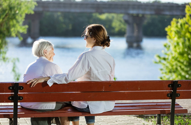 Idős nő és felnőtt unokája ül a padon a nyári parkban és beszél - Fotó, kép